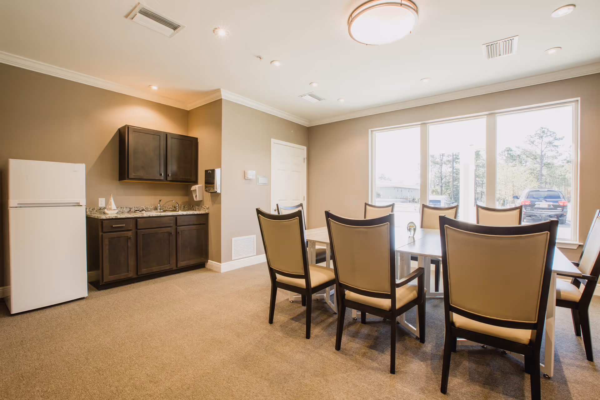 A bright dining room with a rectangular table surrounded by eight beige cushioned chairs with dark wooden frames. The room has beige walls and carpeted floor. On the left side, there is a small kitchenette area with a white refrigerator, dark wooden cabinets, a granite countertop, and a sink. Large windows on the right side let in natural light and show a view of parked cars and trees outside.