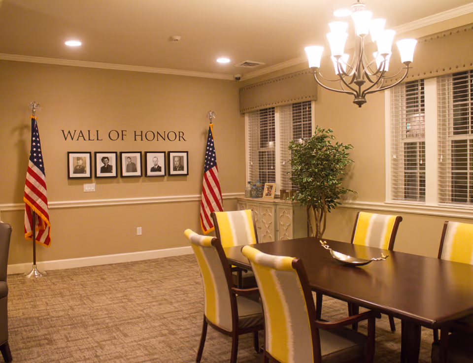 A well-lit room with a dark wooden table surrounded by six chairs with yellow and white upholstery. On the wall, there is a 'Wall of Honor' display featuring five framed black and white portraits, flanked by two American flags. A chandelier hangs above the table, and there is a green potted plant near the windows with white blinds. A decorative cabinet with framed photos is positioned under the windows.