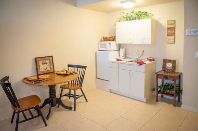 Small kitchenette area with a white mini refrigerator, microwave, and white cabinets. A round wooden table with two wooden chairs is set with plates and napkins. A small wooden side table with a framed picture and a green plant is against the wall. The walls are light-colored and decorated with small framed signs.
