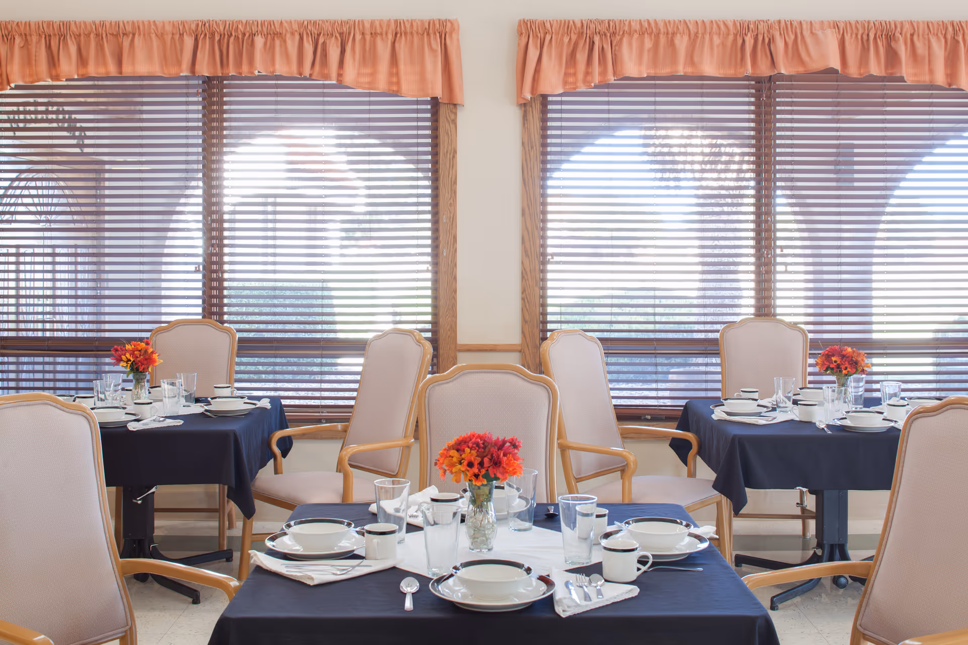 A dining room with several tables covered in dark blue tablecloths, each set with white plates, bowls, cups, silverware, and glasses. Each table has a small vase with red and orange flowers. The room has large windows with wooden blinds and peach-colored valances.