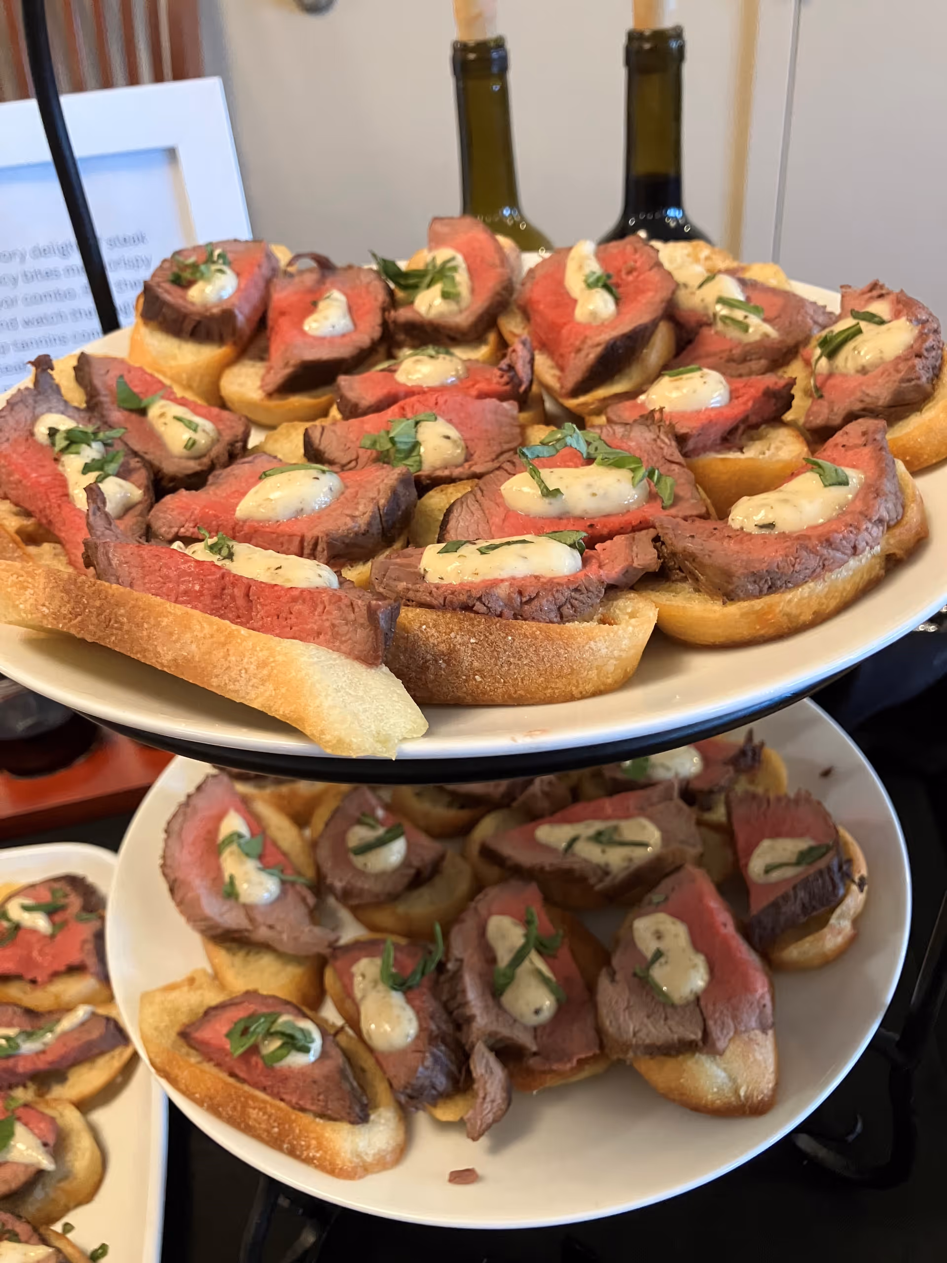 Two-tiered serving platter with slices of toasted baguette topped with medium-rare roast beef, a dollop of creamy sauce, and garnished with fresh herbs. In the background, two bottles of wine are partially visible.