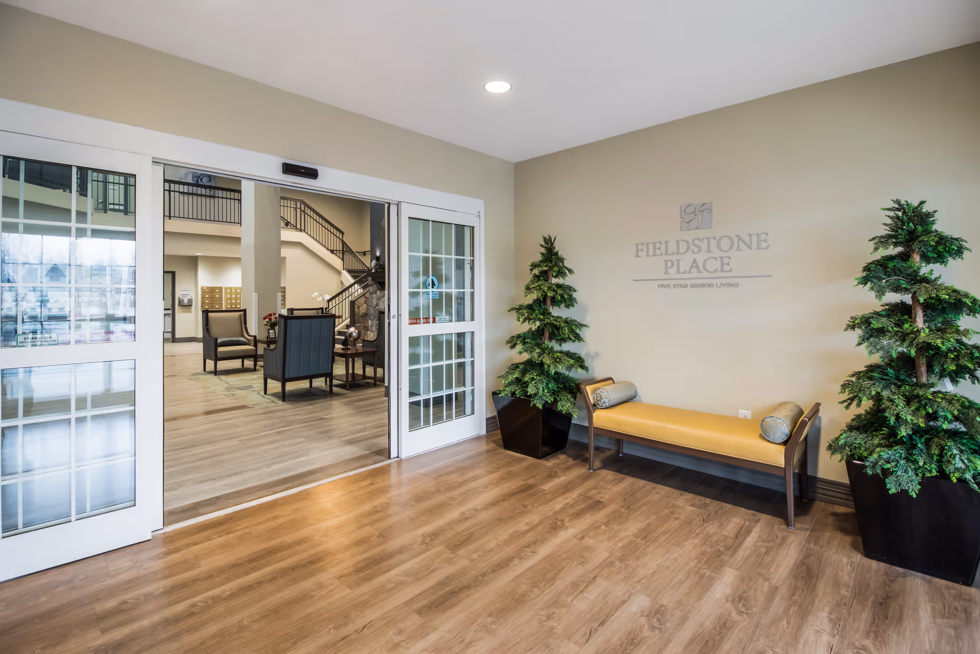 The Fieldstone Place entry lobby with sliding glass doors, a yellow bench flanked by potted plants, and seating visible inside.