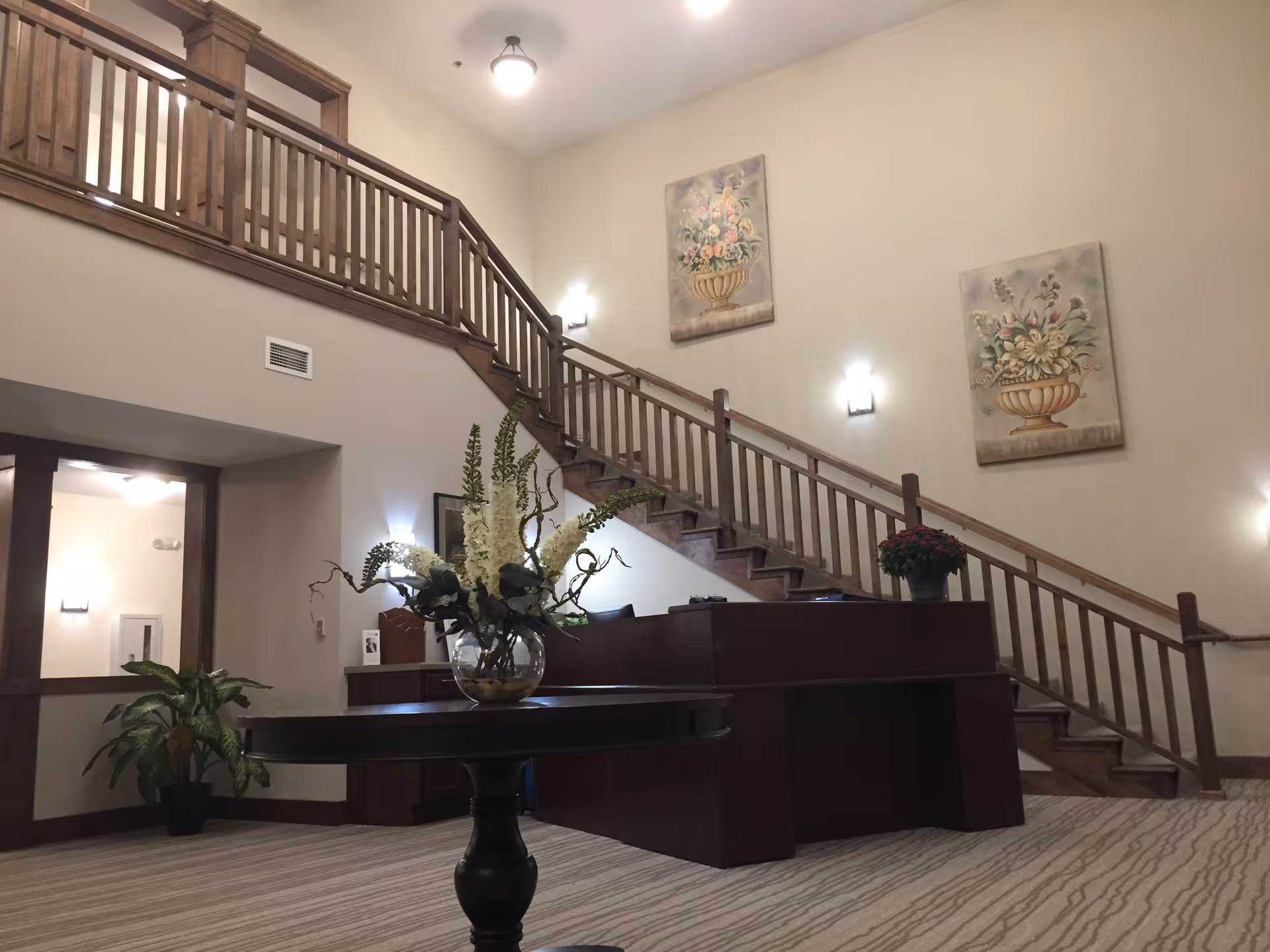 Interior view of a senior living facility lobby with a wooden staircase and railing leading to an upper floor. There is a round table in the foreground with a glass vase holding white flowers and greenery. The walls are decorated with two floral paintings, and there is a reception desk near the stairs. The space is well-lit with ceiling and wall lights, and a potted plant is visible near the entrance.