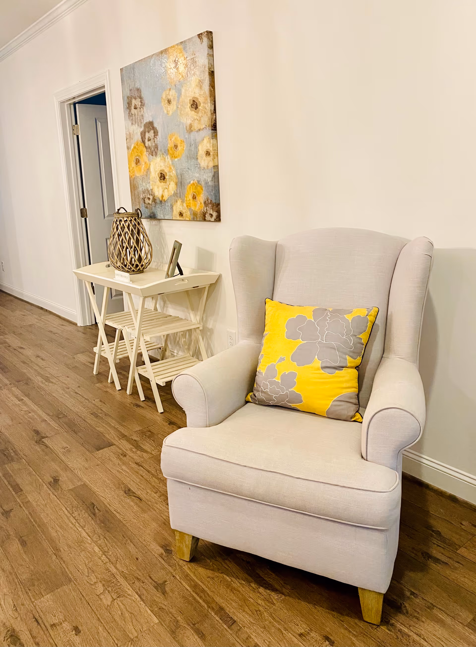 A cozy interior corner featuring a light beige upholstered armchair with a yellow and gray floral cushion. Next to the chair is a small white folding table with a decorative lantern and a small framed photo. Above the table hangs a painting of abstract flowers in shades of yellow, gray, and beige. The floor is wooden, and a white door is visible in the background.