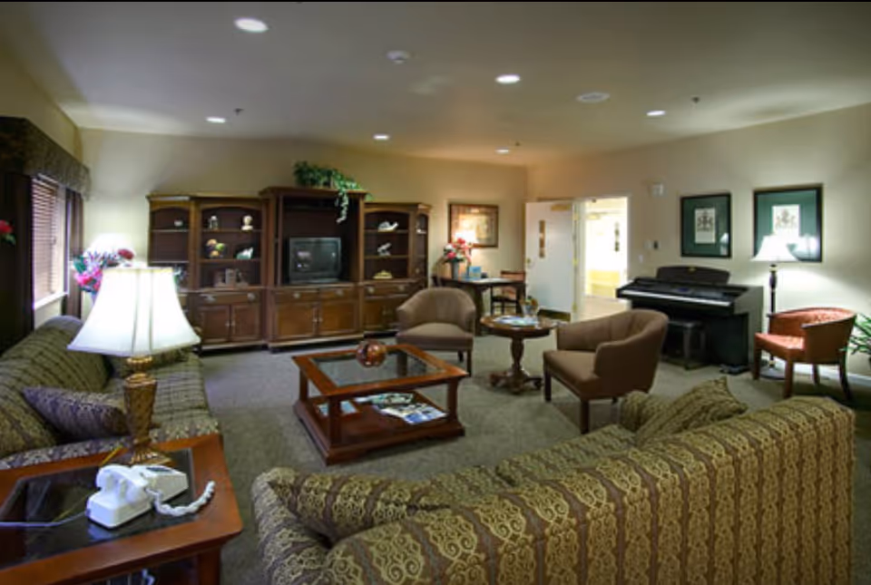 A cozy senior living facility common area with patterned sofas, two armchairs, a wooden coffee table, a TV cabinet, a piano, and a side table with a lamp and telephone. The room is softly lit with ceiling lights and has framed artwork on the walls.