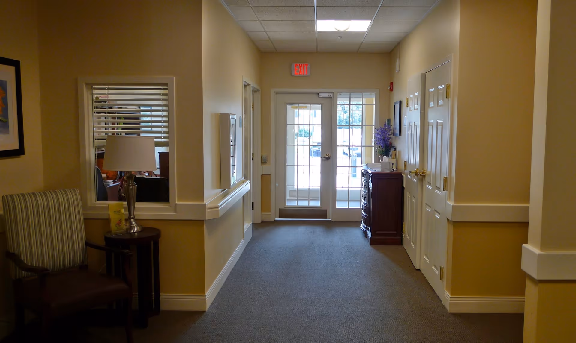Carpeted interior hallway of a senior living facility with chairs and a lamp on the left, a small console with flowers on the right, and glass exit doors at the far end.