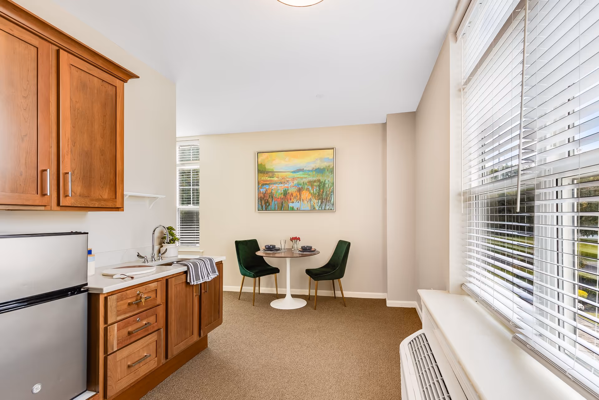 A small dining area with a round table set for two with dark green chairs, next to a kitchenette featuring wooden cabinets, a stainless steel mini fridge, a sink, and a window with blinds letting in natural light. A colorful landscape painting hangs on the wall above the dining table.