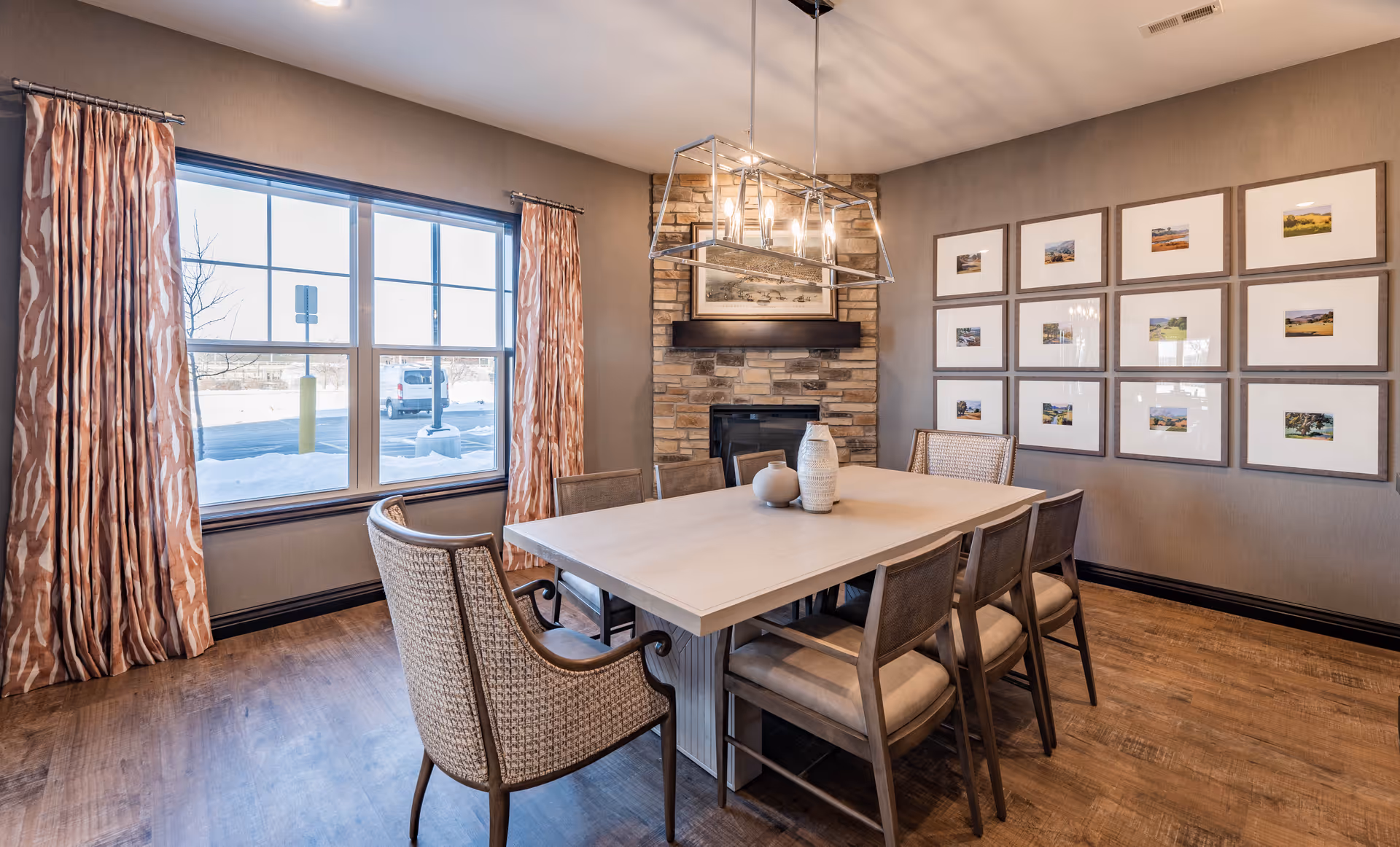 Interior dining room with a rectangular table surrounded by chairs, a stone fireplace, framed artwork on the wall, and a large window with patterned curtains.