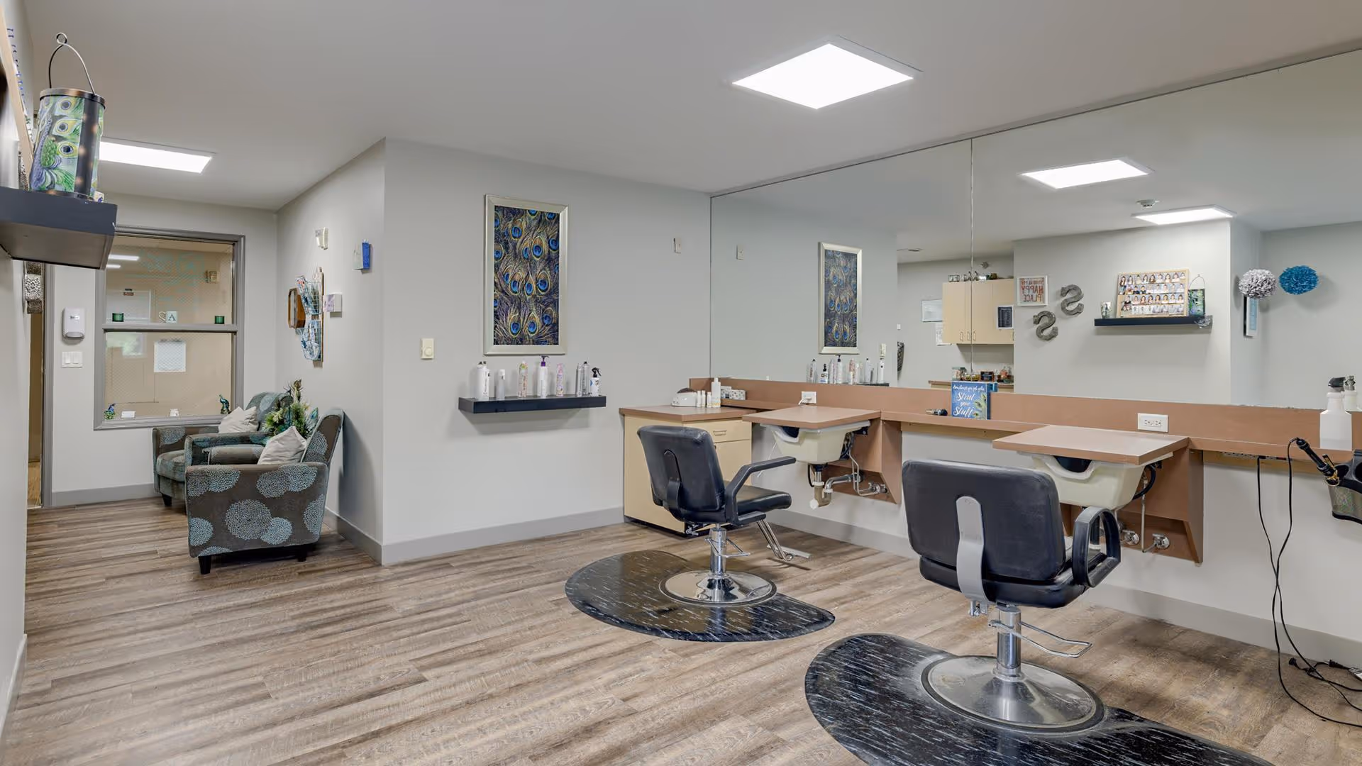 Interior view of a salon area in a senior living facility with two black salon chairs in front of a large mirror and sinks. To the left, there is a seating area with two patterned armchairs and a small table. The room has wood flooring and neutral-colored walls with decorative artwork and shelves holding hair care products.