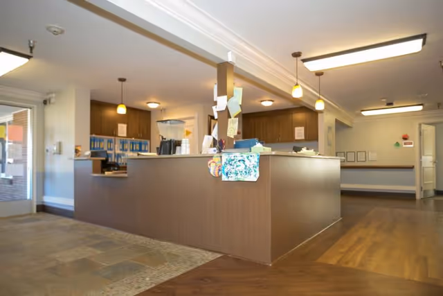 Interior view of a reception or front desk area in a senior living facility with a brown counter, hanging pendant lights, and various papers and decorations attached to the counter. The floor is a combination of tile and wood, and there are doors and bulletin boards visible in the background.