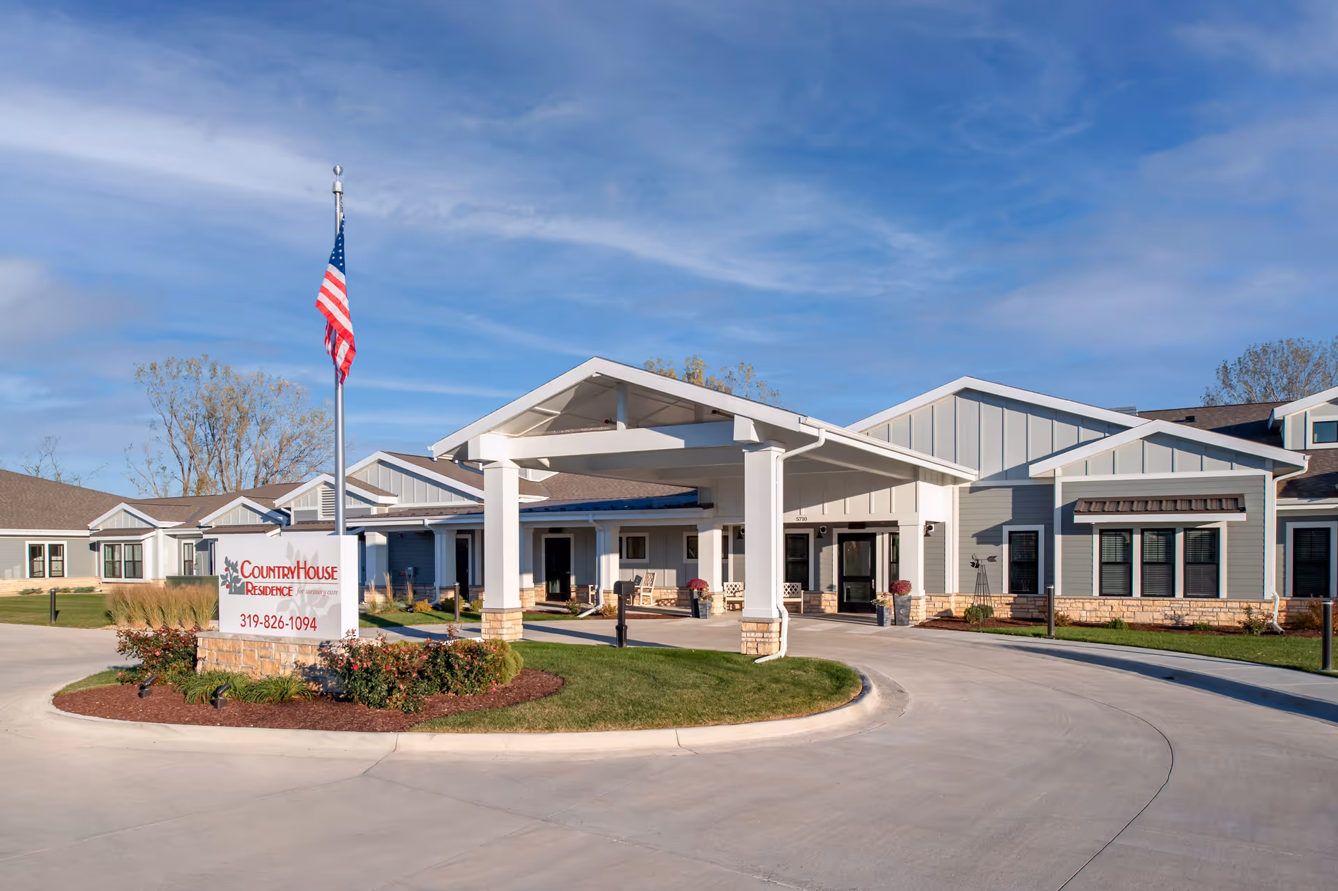 Exterior view of CountryHouse Residence, a single-story senior living facility with a covered entrance, a circular driveway, and an American flag on a flagpole. The building has light gray siding with stone accents and several windows. There is a landscaped area with flowers and shrubs around the sign displaying the facility's name and phone number.