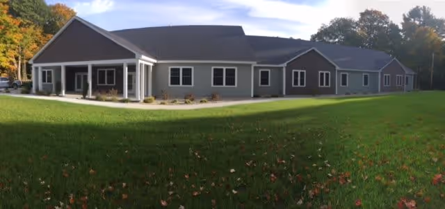 Wide exterior view of a single-story assisted living facility building with gray siding, white trim, and a dark roof, surrounded by green grass and trees with autumn foliage under a partly cloudy sky.