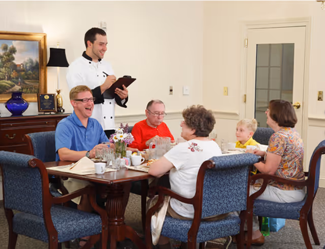 A group of five people, including an elderly man, two women, a young boy, and a man in a blue shirt, are seated around a dining table in a well-lit room. A waiter in a white uniform is standing beside the table, taking their order on a clipboard. The table is set with cups, glasses, and a small flower arrangement. The room has traditional decor with a painting, a lamp, and a wooden sideboard in the background.