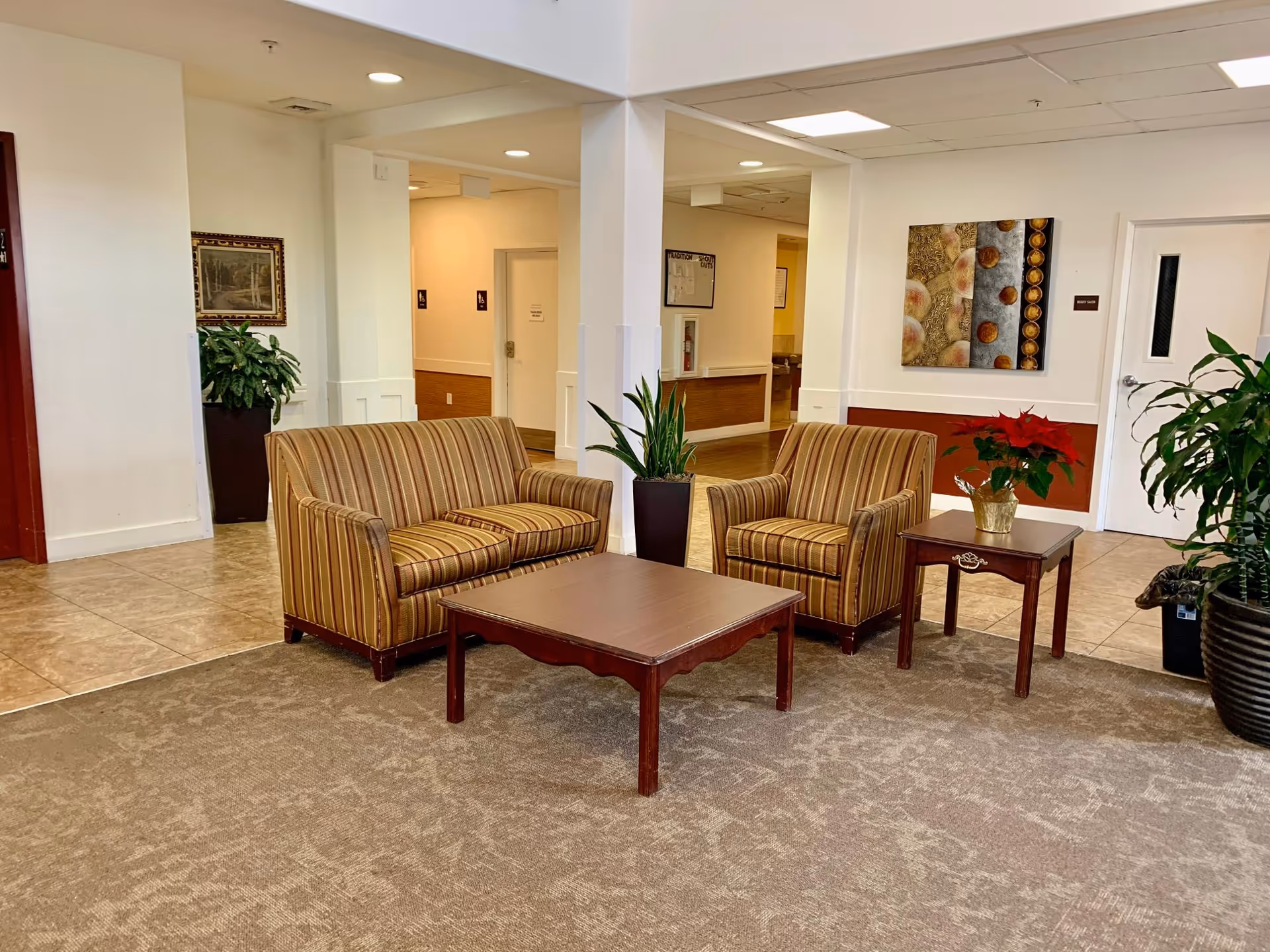 A cozy seating area in an assisted living facility with a striped loveseat and armchair, a wooden coffee table, and a side table with a potted poinsettia plant. The area is carpeted and surrounded by tiled flooring, with potted plants and framed artwork on the walls. The space is well-lit with ceiling lights and has white walls with a reddish-brown accent panel.