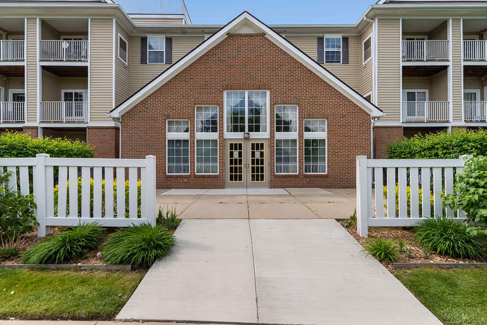 Front exterior view of Elmhaven Manor featuring a brick building with large windows and double glass doors, flanked by beige siding and balconies. A concrete pathway leads to the entrance, bordered by white fences and green shrubs.