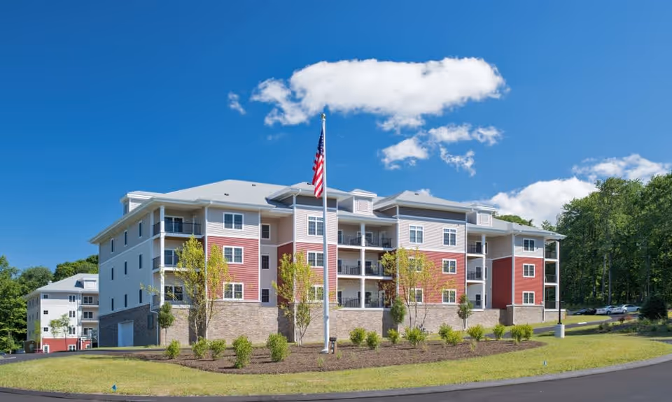 Exterior view of a multi-story senior living facility building with red and white siding, balconies, and an American flag on a flagpole in front, surrounded by landscaped greenery and trees under a blue sky with some clouds.