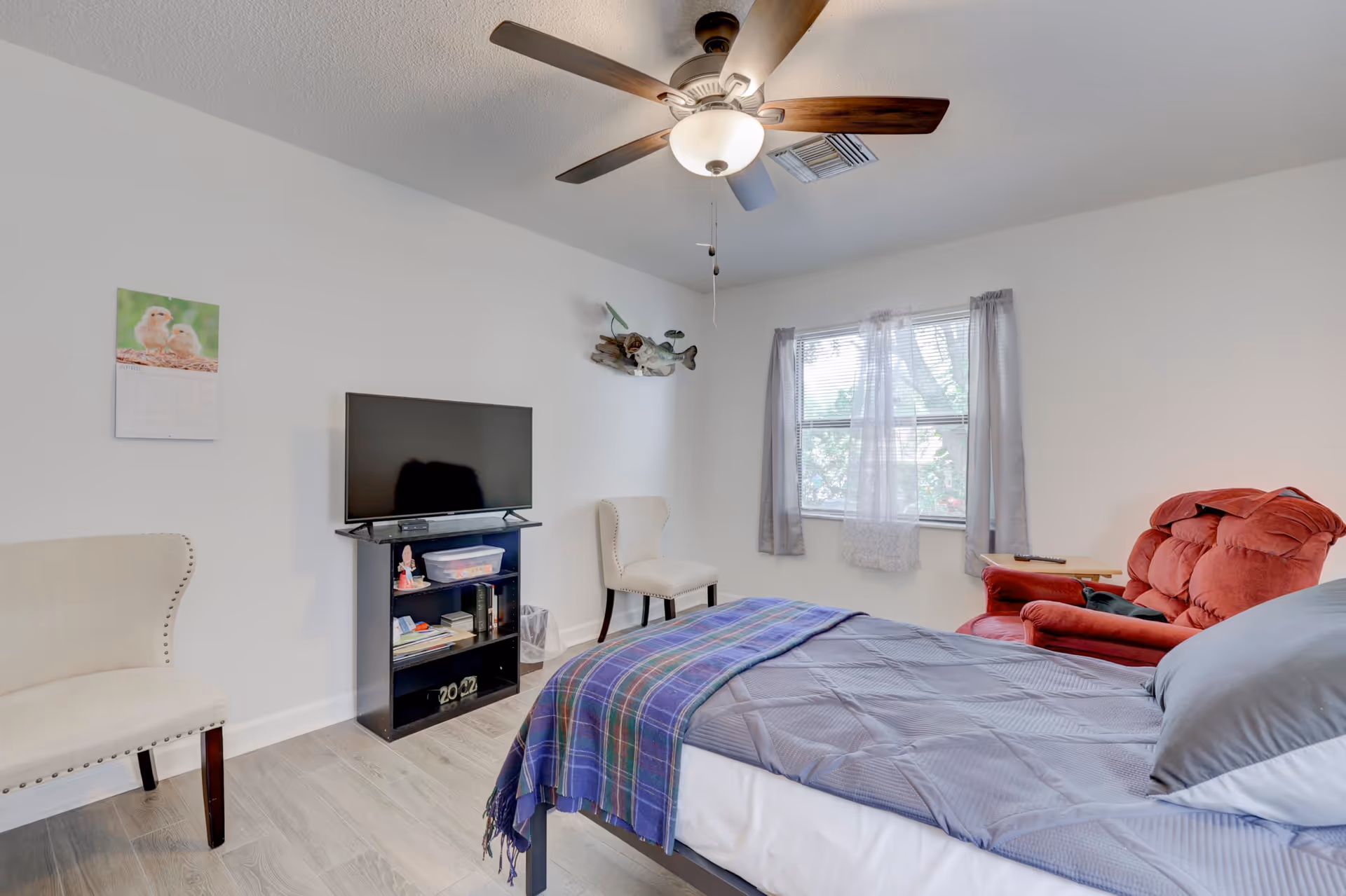 A bright bedroom with a bed covered in gray bedding and a purple plaid blanket. There is a red recliner chair next to a window with sheer curtains, two beige chairs, a black TV stand with a flat-screen TV, and a ceiling fan with a light fixture.