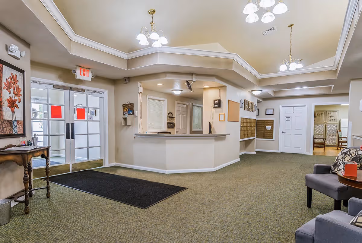 Interior view of a senior living facility lobby area with a reception desk, green carpet, beige walls, ceiling lights, a small table with a flower painting on the left, and seating area with chairs on the right. Double glass doors with exit sign are visible in the background.