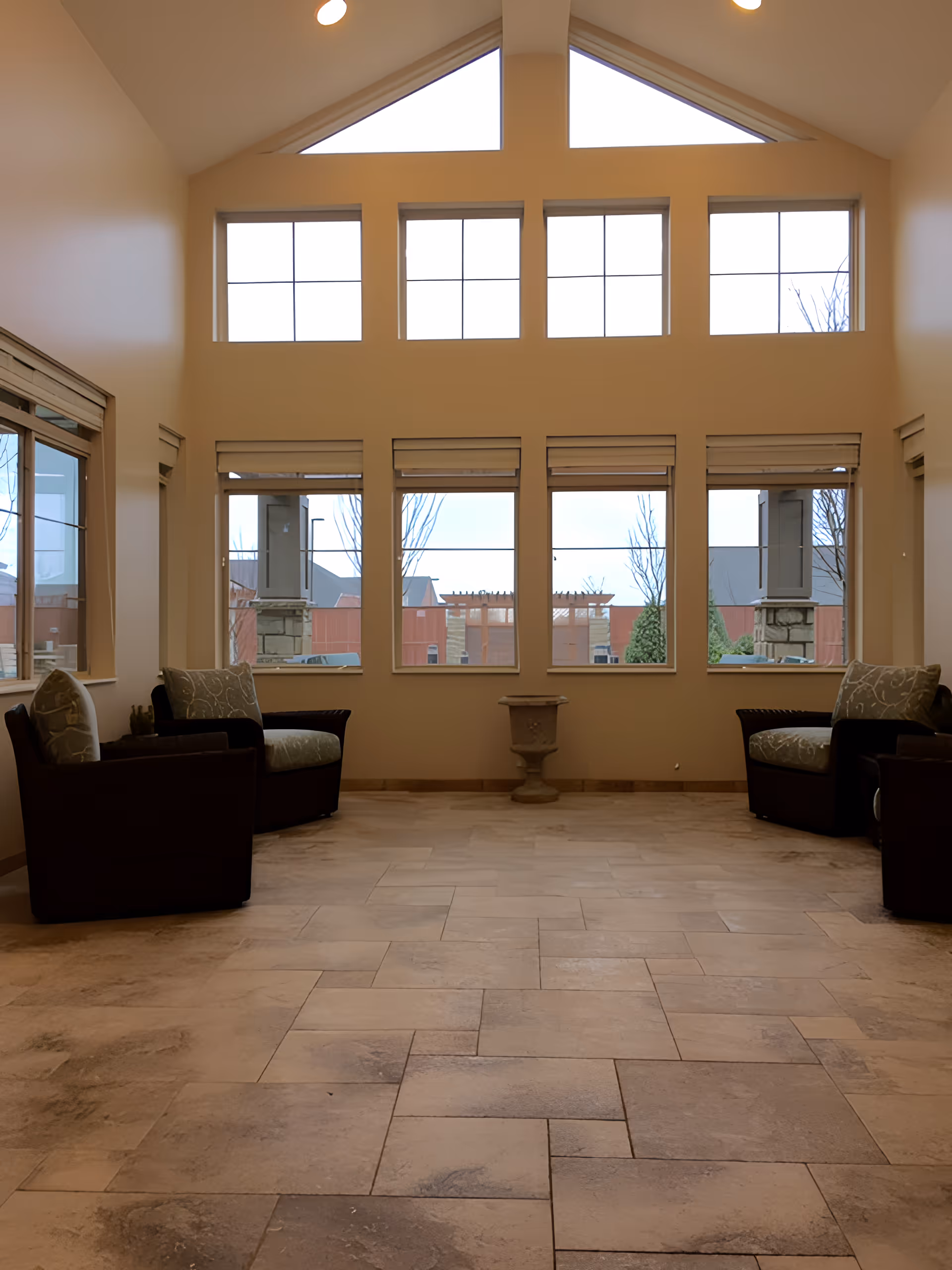 Sunlit high-ceiling common room with large grid windows, tiled floor and armchairs along the sides.