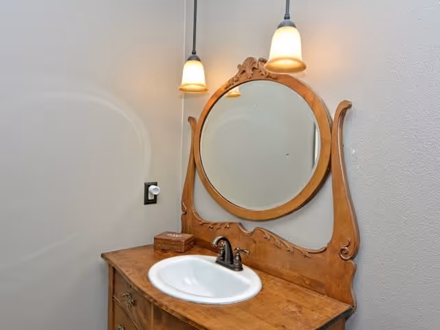 A bathroom vanity with a wooden countertop and an ornate wooden framed round mirror. There is a white sink with a dark faucet, a small wooden box on the counter, and two pendant lights hanging above the mirror. The walls are painted light gray.