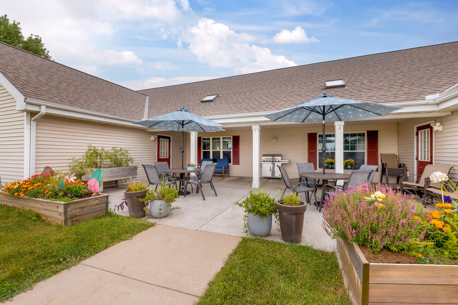 Outdoor patio area at Brookdale Willmar with tables and chairs under two large umbrellas. There are several potted plants and raised garden beds with colorful flowers surrounding the concrete patio. The building has beige siding with red doors and windows, and a grill is visible on the patio.