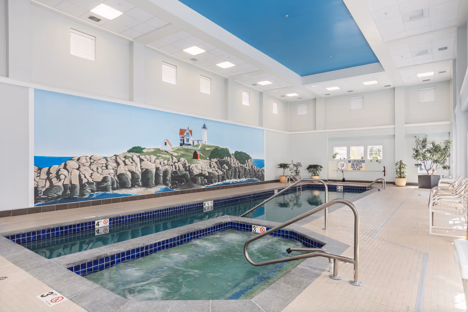 Bright indoor swimming pool and adjacent hot tub in a high-ceiling room with a lighthouse mural on the wall.