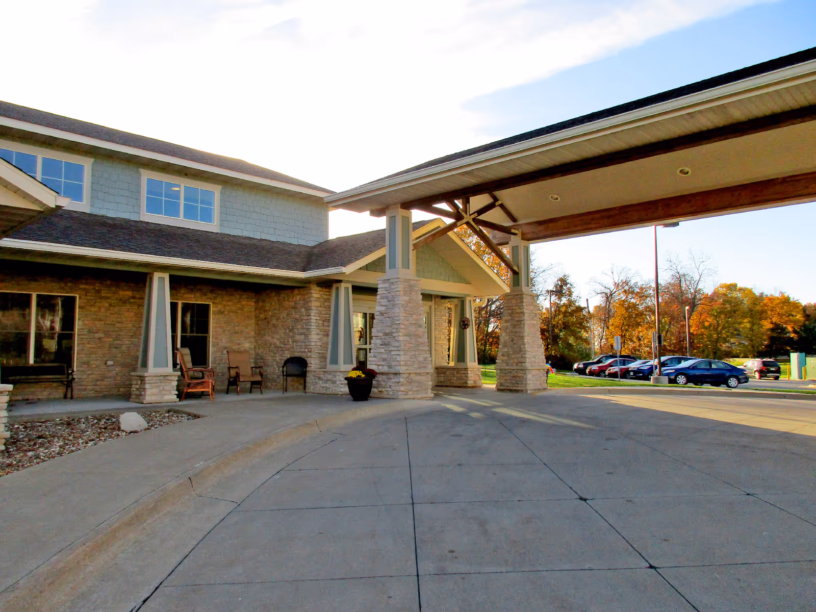 Exterior view of the entrance to Good Samaritan Society – Ottumwa, showing a covered driveway with stone pillars, a seating area with chairs, and a parking lot with cars in the background under a partly cloudy sky.