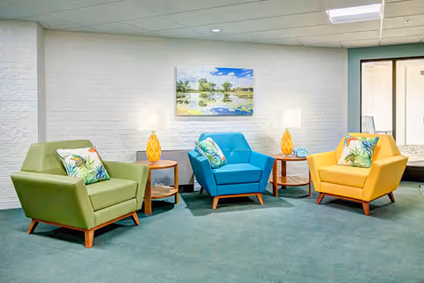 Three colorful armchairs (green, blue, and yellow) arranged with side tables and lamps against a white brick wall beneath a landscape painting.
