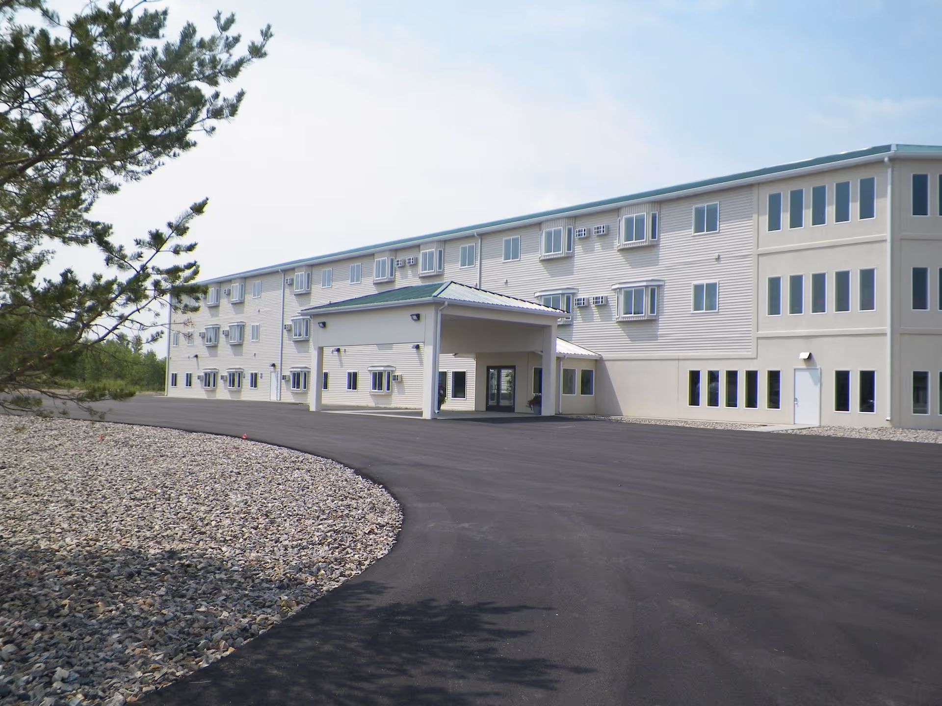Exterior view of a three-story senior living facility building with multiple windows and a covered entrance. The building is surrounded by a paved driveway and landscaping with rocks and a tree on the left side.