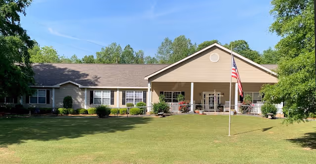 Single-story beige building with a gable roof, surrounded by green trees and bushes, with a large grassy lawn in front and an American flag on a flagpole near the entrance.