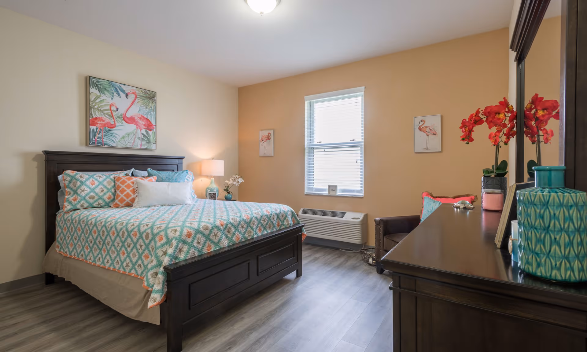 A cozy bedroom with a dark wooden bed frame and a colorful patterned bedspread in shades of teal, orange, and white. Above the bed is a painting of two pink flamingos. There is a window with blinds letting in natural light, a small armchair with a teal pillow, and a dark wooden dresser with a mirror. On the dresser are decorative items including a vase with red flowers and a teal ceramic container. The walls are painted a warm beige color and the floor has light wood laminate.