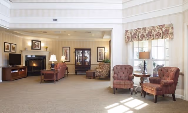 A spacious and well-lit living room area with beige walls and carpet. The room features two upholstered armchairs with a small round table and lamp between them near a window with floral valance. In the background, there is a seating area with a sofa, armchair, coffee table, lamps, a fireplace, framed artwork on the walls, and a wooden cabinet.
