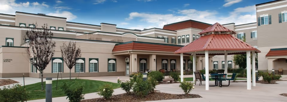 Outdoor view of a senior living facility with a beige multi-story building in the background, featuring multiple windows and a red-roofed gazebo with seating in the foreground. There are landscaped areas with small trees, bushes, and a paved walkway.