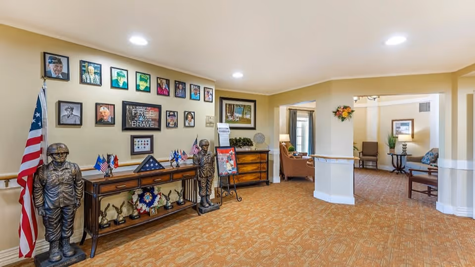 Interior view of a senior living facility hallway with a military tribute display including statues of soldiers, an American flag, framed photos, and patriotic decorations. The hallway leads to a seating area with chairs and a table, decorated with plants and artwork.