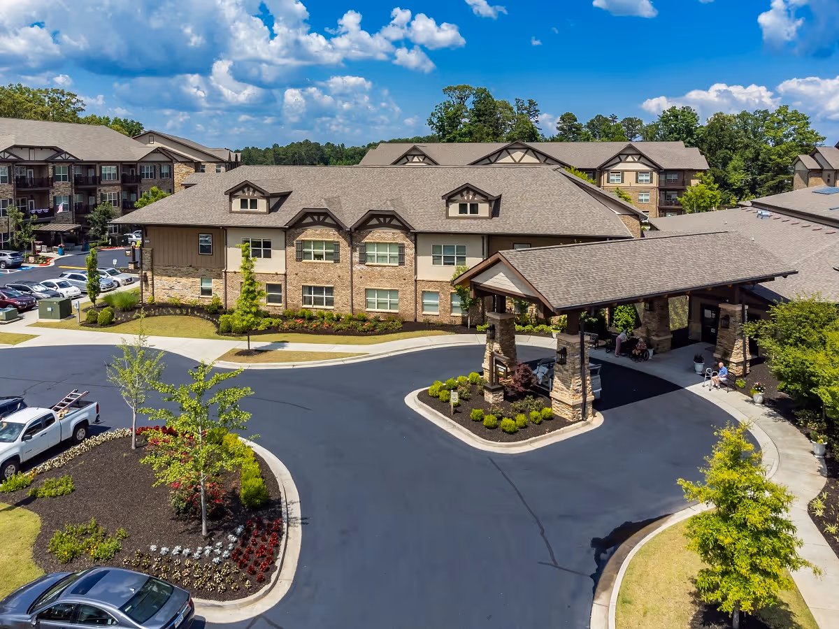 Exterior view of Celebration Village Forsyth senior living facility showing a large building with multiple windows, a covered entrance with stone pillars, landscaped gardens, parked cars, and a bright blue sky with scattered clouds.