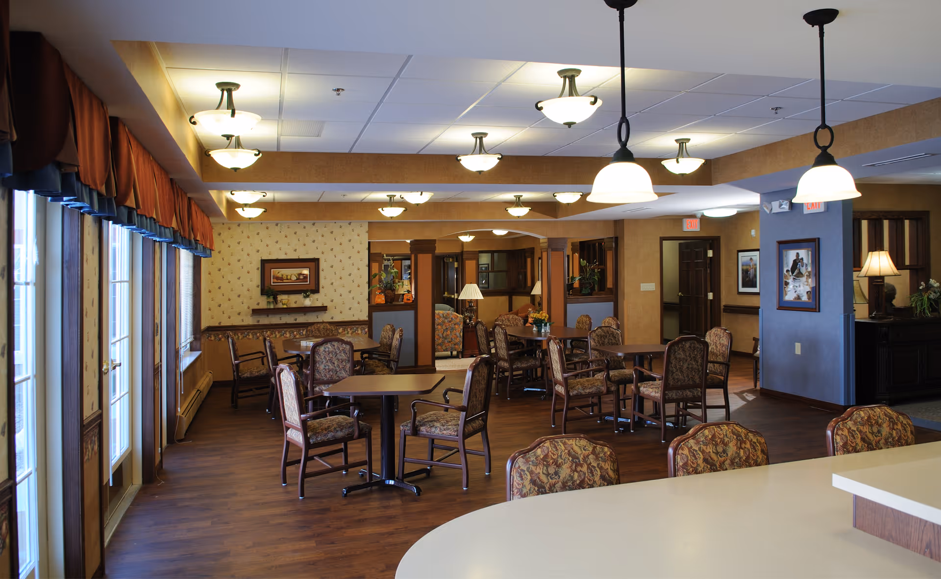 A spacious dining room in a senior living facility with multiple tables and chairs arranged neatly. The room features wooden flooring, warm beige walls with framed artwork, and several ceiling lights providing ample illumination. Large windows with red curtains allow natural light to enter the room. A countertop with pendant lights is visible in the foreground.