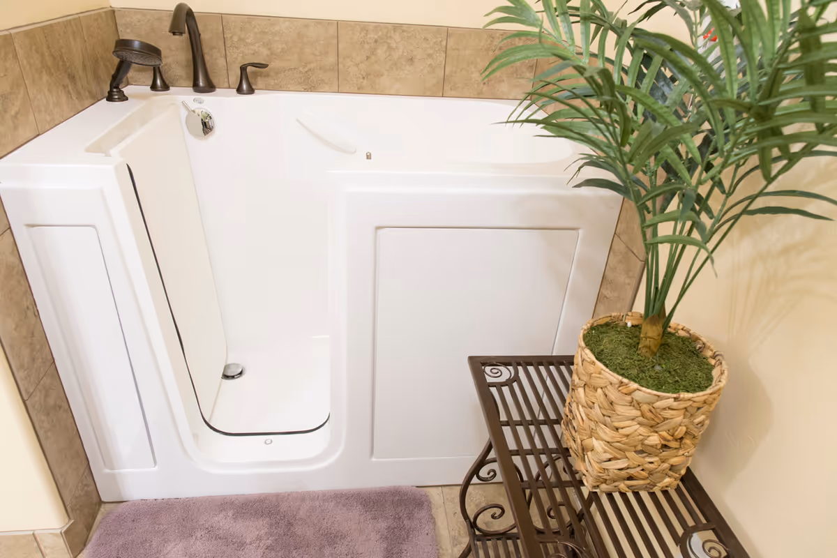 A white walk-in bathtub with a door and bronze faucet fixtures, surrounded by beige tiled walls. Next to the tub is a metal rack holding a woven basket with a green leafy plant. A purple bath mat is placed on the floor in front of the tub.