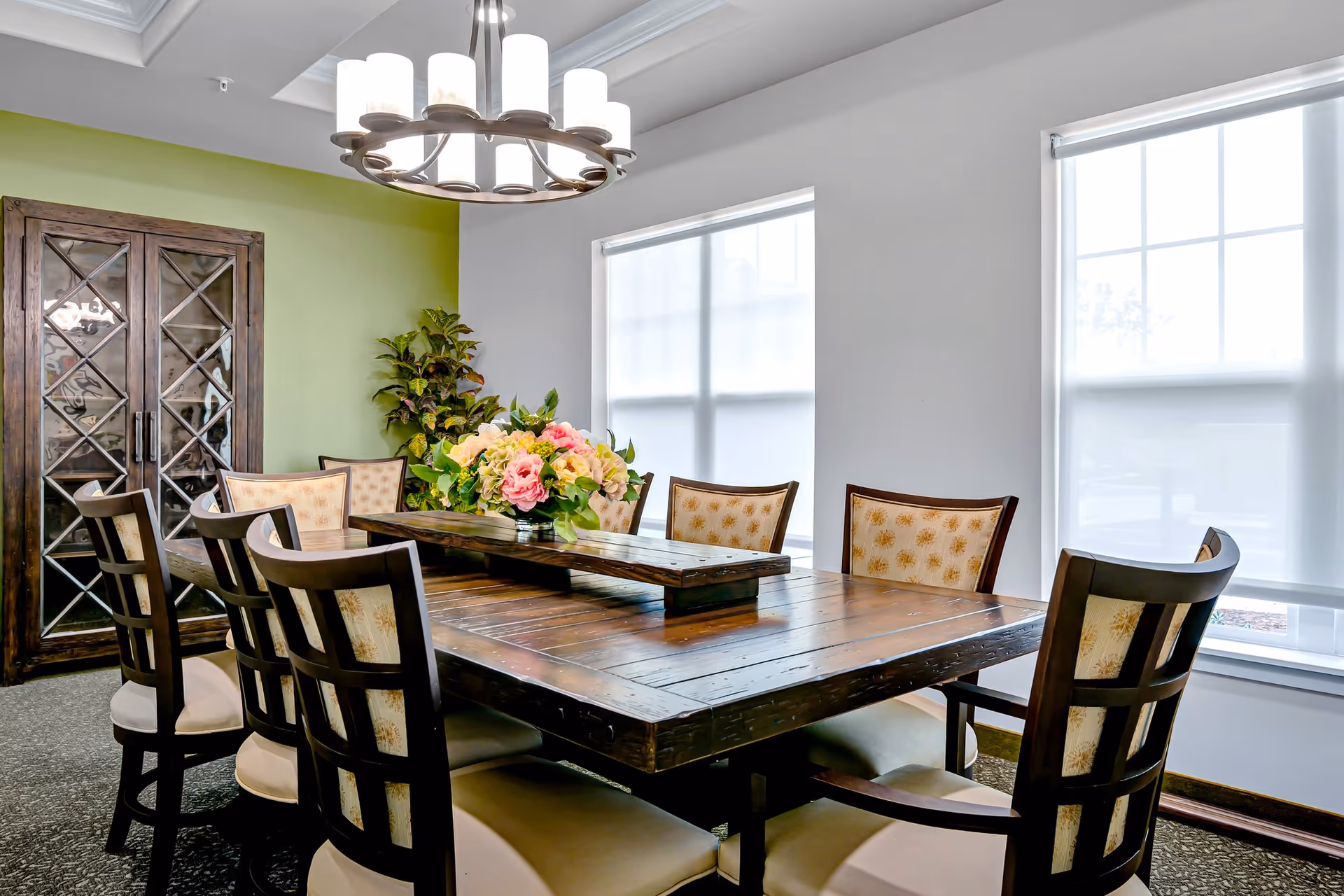 A bright dining room with a large wooden table surrounded by eight cushioned chairs. The table has a floral centerpiece with pink and yellow flowers. The room features two large windows with white blinds, a green accent wall, a wooden cabinet with glass doors, and a modern chandelier hanging above the table.