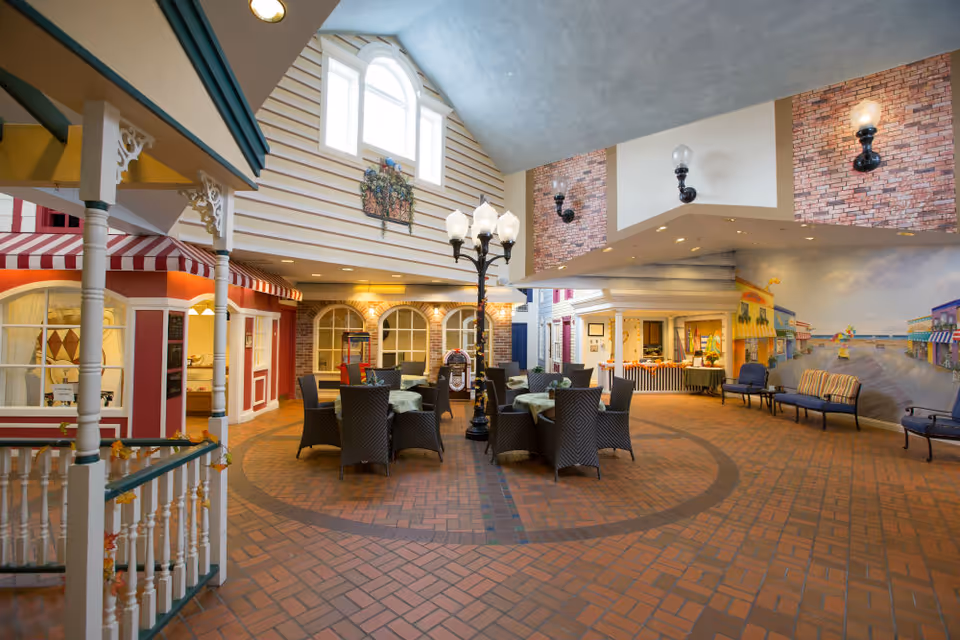 Indoor community atrium with round dining tables and wicker chairs, a central streetlamp, and storefront-style walls and murals.