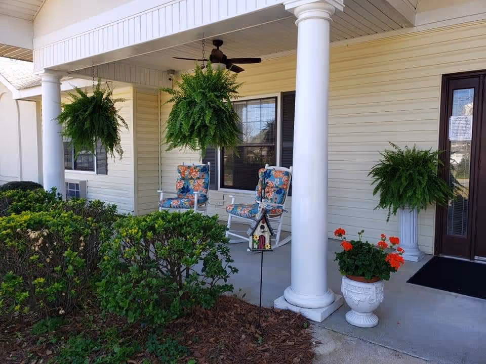 Front porch area of a building with white columns, two floral cushioned rocking chairs, hanging green ferns, a potted plant with red flowers, and a small decorative birdhouse on a stake in the garden bed.