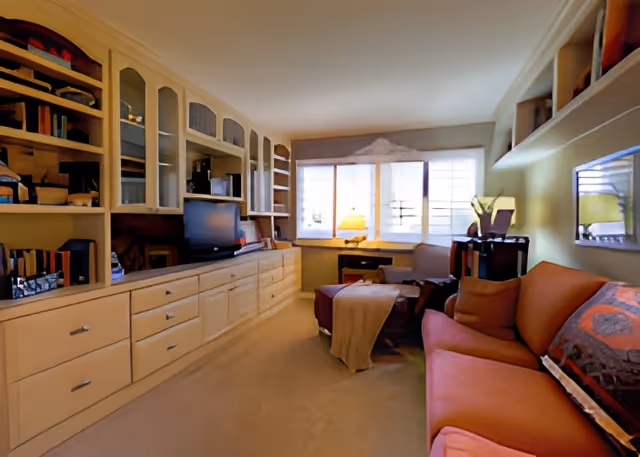 A cozy living room with a long built-in cabinet and shelves on the left wall, housing a television and various decorative items. On the right side, there is an orange sofa with cushions and a blanket, a small table with a lamp, and a window with blinds letting in natural light.