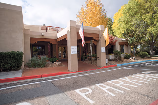 Exterior view of the entrance to Brookdale Santa Fe facility with two flags displayed on pillars, surrounded by trees with autumn foliage and a parking area in front.