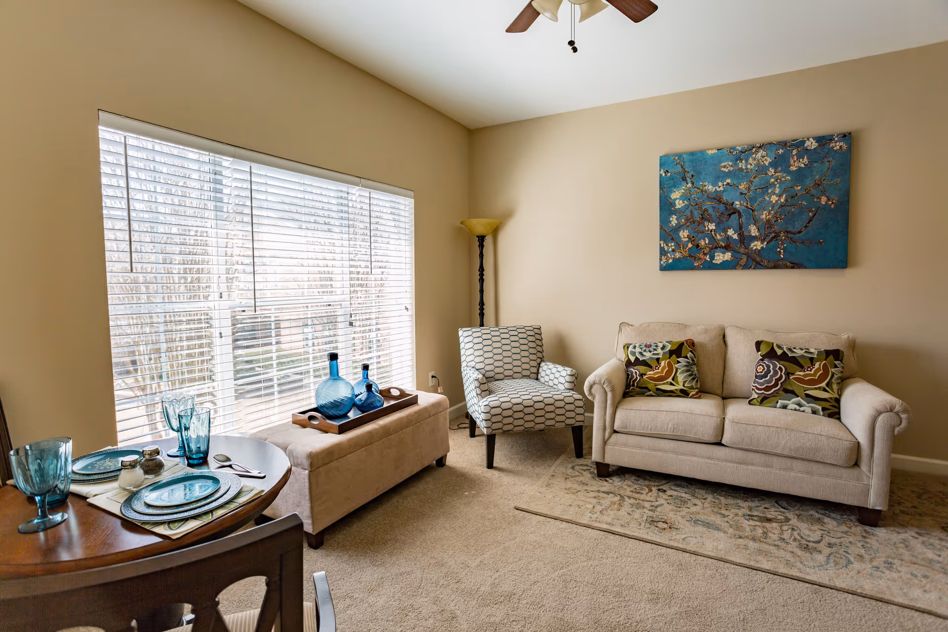 A cozy living room with a beige sofa adorned with patterned cushions, a patterned armchair, a floor lamp, and a large window with white blinds. A small round dining table is set with blue glassware and plates. A decorative tray with blue glass bottles sits on a beige ottoman. A blue floral painting hangs on the wall above the sofa.