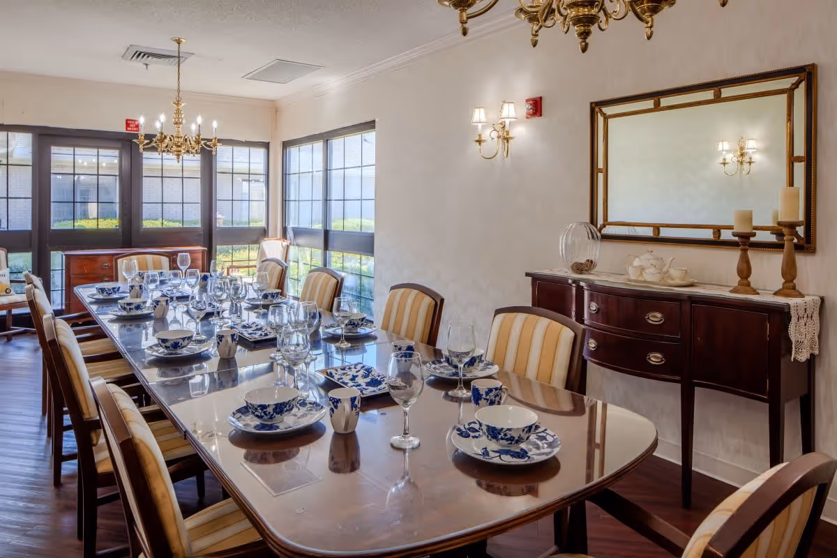 A formal dining room with a long wooden table set with blue and white patterned dishes, cups, and glassware. The room has striped upholstered chairs around the table, large windows letting in natural light, a chandelier hanging from the ceiling, and a sideboard with candles and a mirror above it.