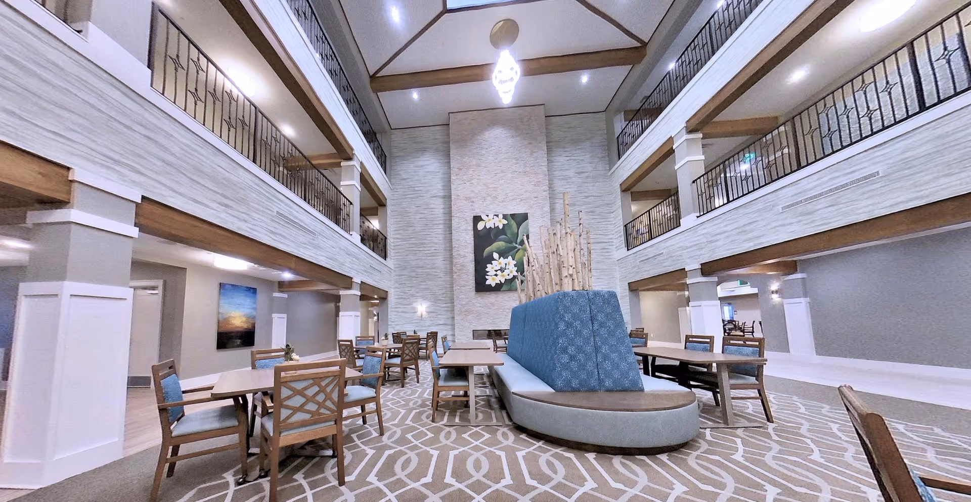 Spacious and well-lit common area in a senior living facility with high ceilings and two levels of balconies. The room features multiple wooden tables and chairs with blue cushions, a central curved bench with blue upholstery, a patterned carpet, and a large floral painting on the far wall. The walls are light-colored with white pillars and wooden accents.