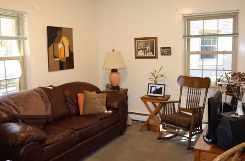 Cozy living room with a brown leather sofa, wooden rocking chair, side table with a lamp and laptop, framed wall art and two windows.