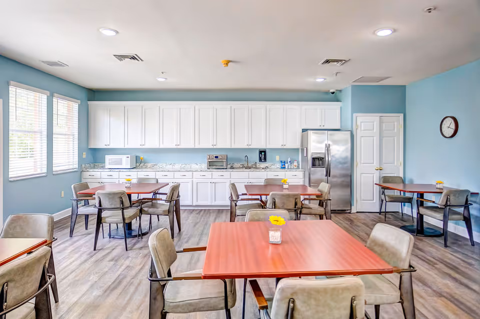 A bright dining room with light blue walls and wood flooring featuring several square wooden tables with gray cushioned chairs. The back wall has white cabinets, a microwave, a toaster oven, a sink, and a stainless steel refrigerator. There are windows on the left side letting in natural light and a clock on the right wall.