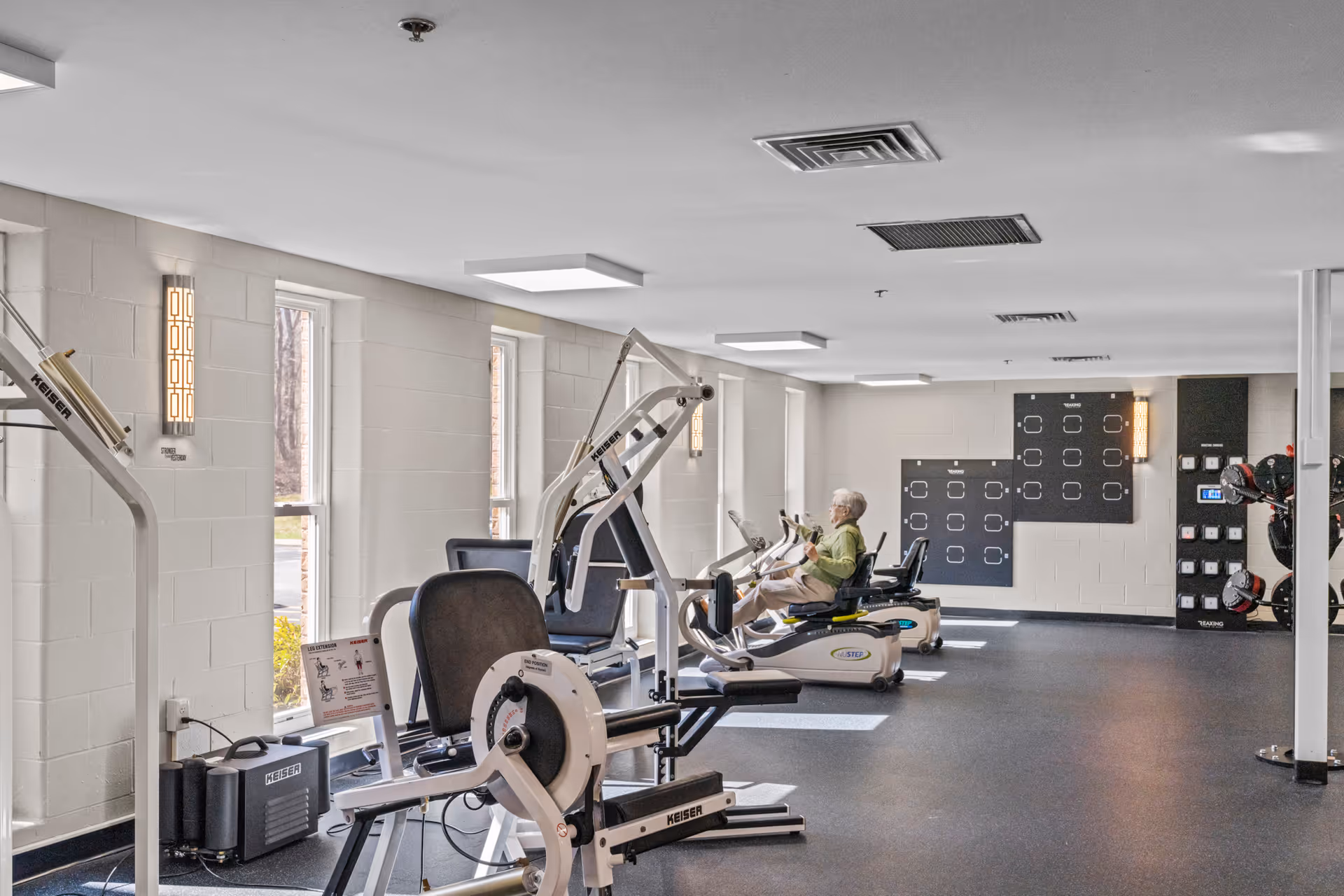 A senior woman exercising on a recumbent bike in a well-lit fitness room with various exercise machines and large windows along one wall.