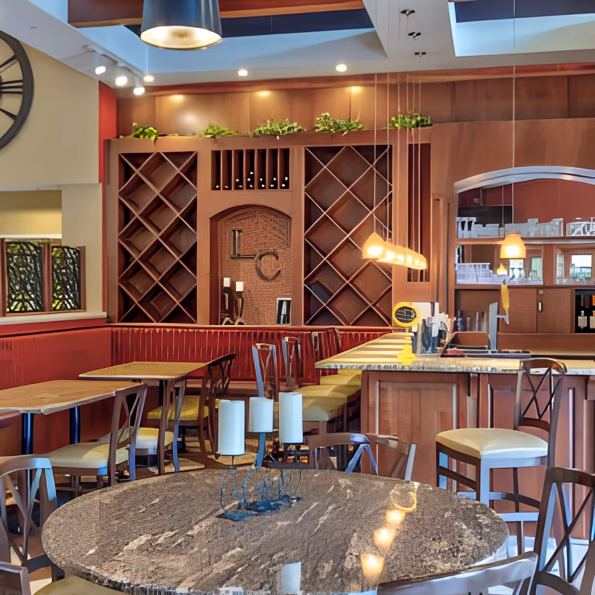 Interior view of a dining area with wooden tables and chairs, a granite round table with a candle holder centerpiece, a bar counter with high chairs, wine racks on the wall, and hanging pendant lights providing warm illumination.