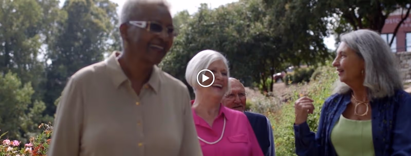 A group of four older adults walking outdoors along a garden path, smiling and engaging in conversation with trees and greenery in the background.